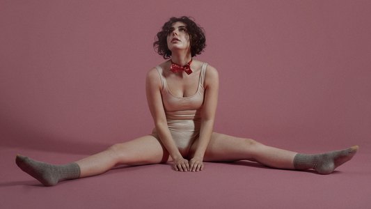 Crazy Moments in Vaginal Health History - Model in Beige Leotard and Grey Socks Sitting with Pink Background 