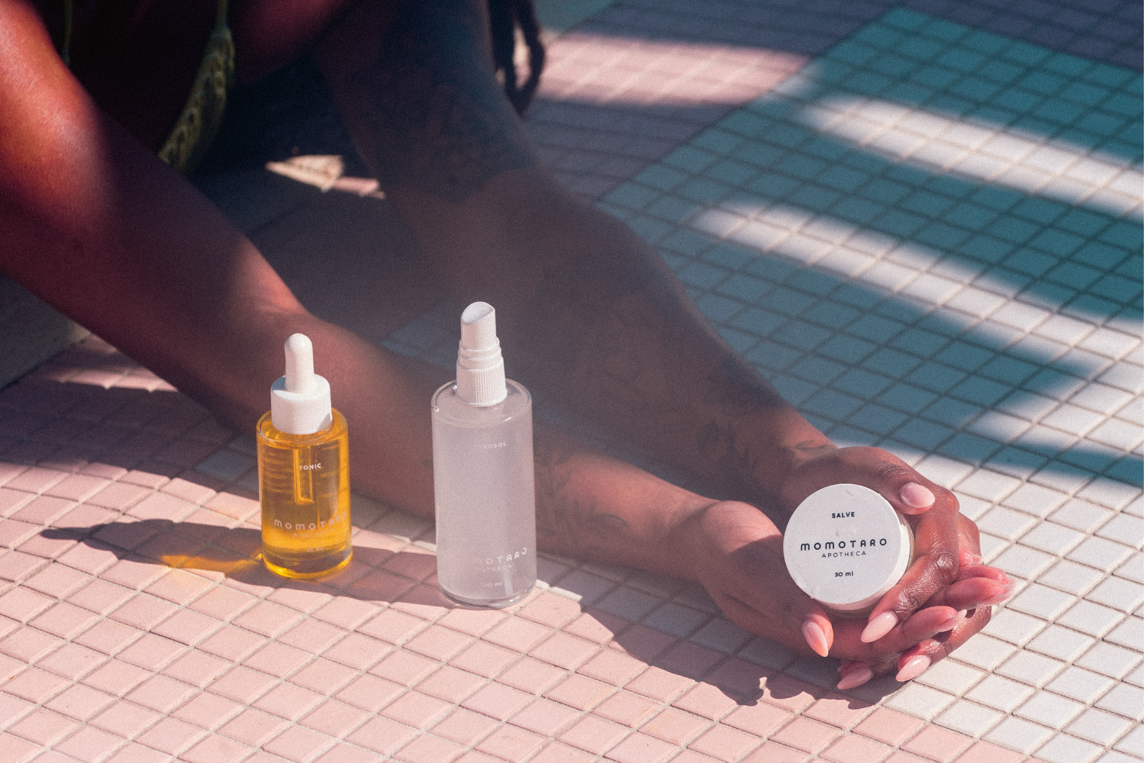A person's tattooed arms and hands rest on a tiled floor next to three momotaro apotheca products. 